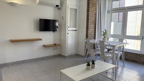 Ett modernt, väl upplyst rum med en platt-TV som är monterad på väggen, ett litet bord med stolar och ett fönster med persienner.