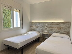 Een kamer met twee eenpersoonsbedden, een houten hoofdbord, een klein nachtkastje en een raam met uitzicht op bomen.
