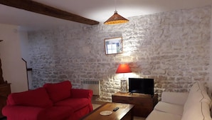 A living room with a red sofa, a white sofa, a wooden coffee table, a TV on a wooden stand, a lamp, and a stone wall. - Two Bedroom House in Domaine les Granges (Ternant)