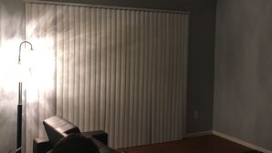 A room with a large window covered by blinds, a floor lamp, and a chair.
