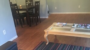 A living room with a dark brown sofa, a wooden coffee table, and a striped rug. There is a dining area with a table and chairs in the background.