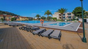 A pool area with sun loungers and a lifebuoy, surrounded by buildings and palm trees.