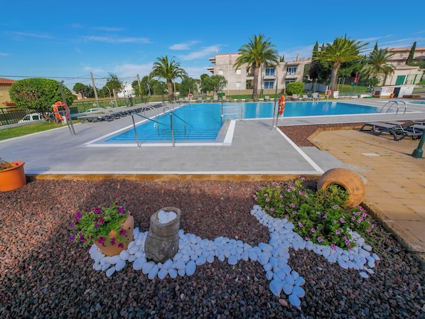 A swimming pool with a concrete deck, surrounded by pebbles and plants, with palm trees and buildings in the background.