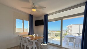 A room with a dining table, chairs, a ceiling fan, a TV, and a large sliding glass door leading to an outdoor balcony. - Agi Rescator 104 Apartamento for rent in Roses Girona. (Rosas / Roses)