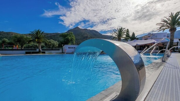 Une piscine dotée d’un élément décoratif aquatique aux formes arrondies, entourée de palmiers et de bâtiments.