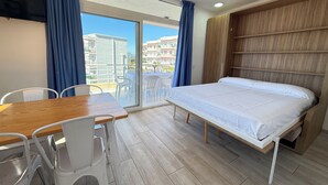 Une pièce comprenant un lit, une table, des chaises, un téléviseur et une vue sur les bâtiments, visible à travers une porte vitrée.
