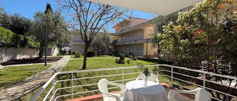 A terrace with white plastic chairs and a table, surrounded by a white railing, with a view of a building and a garden.