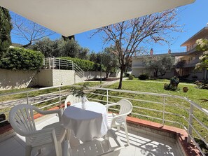 An outdoor patio with a table and chairs, surrounded by a white railing, with a clear view of the courtyard and buildings in the background.