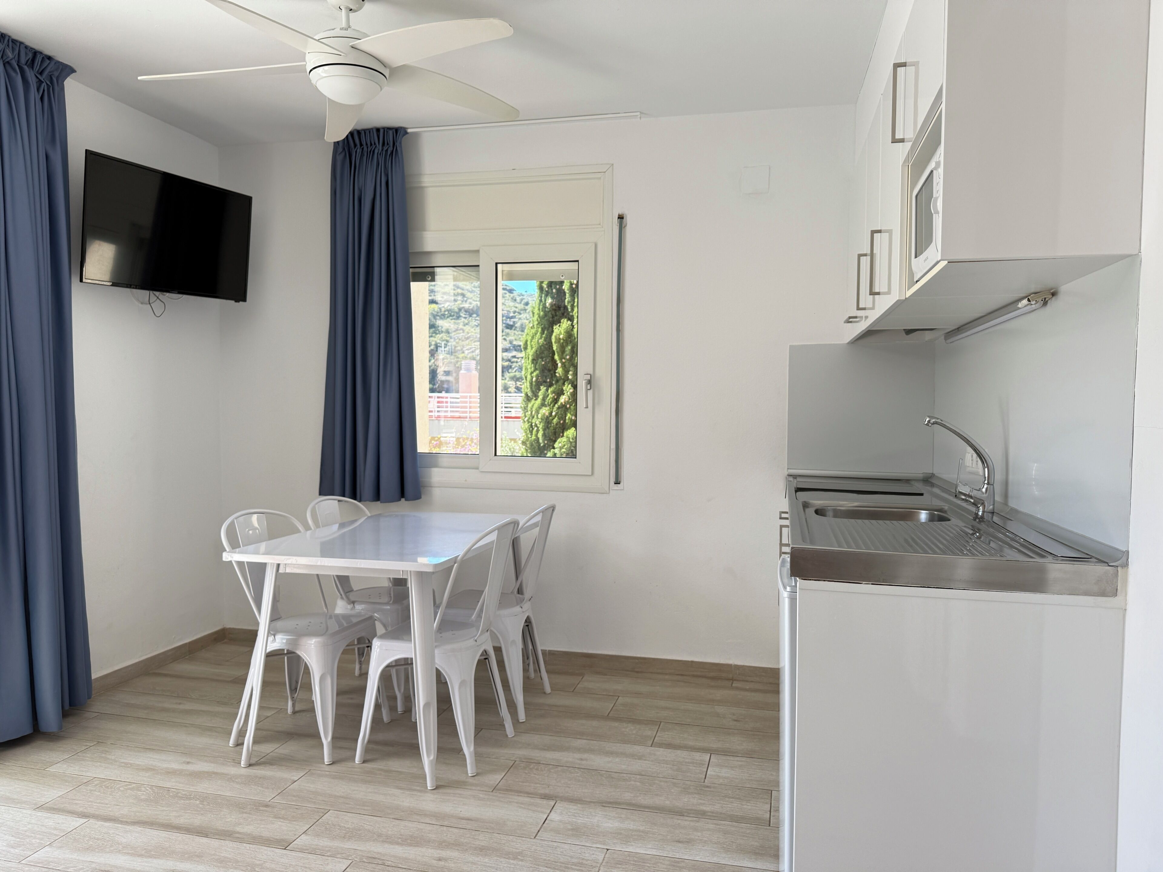 Eine kleine, moderne Küche mit Esstisch und Stühlen, einem Deckenventilator, einem Flachbildfernseher und einem Fenster mit Vorhängen.