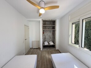 A room with a ceiling fan, a wooden floor, a white bed, a white bench, a radiator, a wooden wardrobe, and a window with a view of trees.