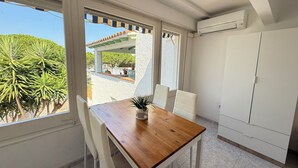 A room with a wooden table and white chairs, large windows with a view of greenery and a building outside, and a white wardrobe.