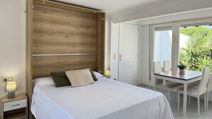 A bedroom with a large bed, bedside table with a lamp, a desk with chairs, and a view of greenery outside.
