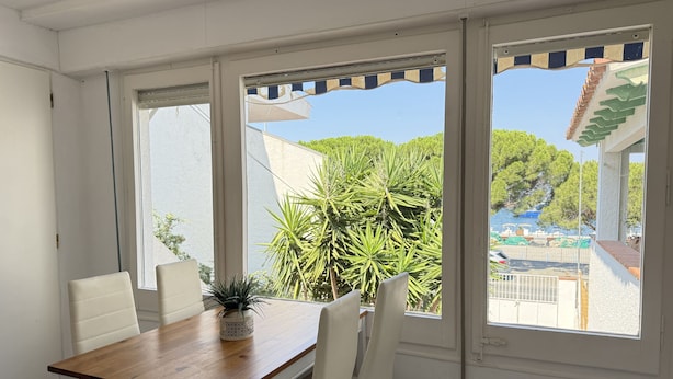 A dining area with a wooden table and white chairs, large windows with a view of greenery and a building with a tiled roof.