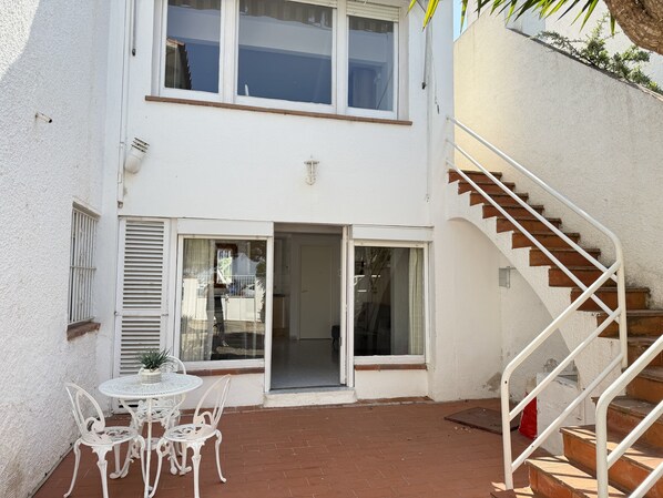 Un edificio de dos pisos con un patio que incluye una mesa y sillas blancas, una escalera y una puerta que da a un espacio interior.