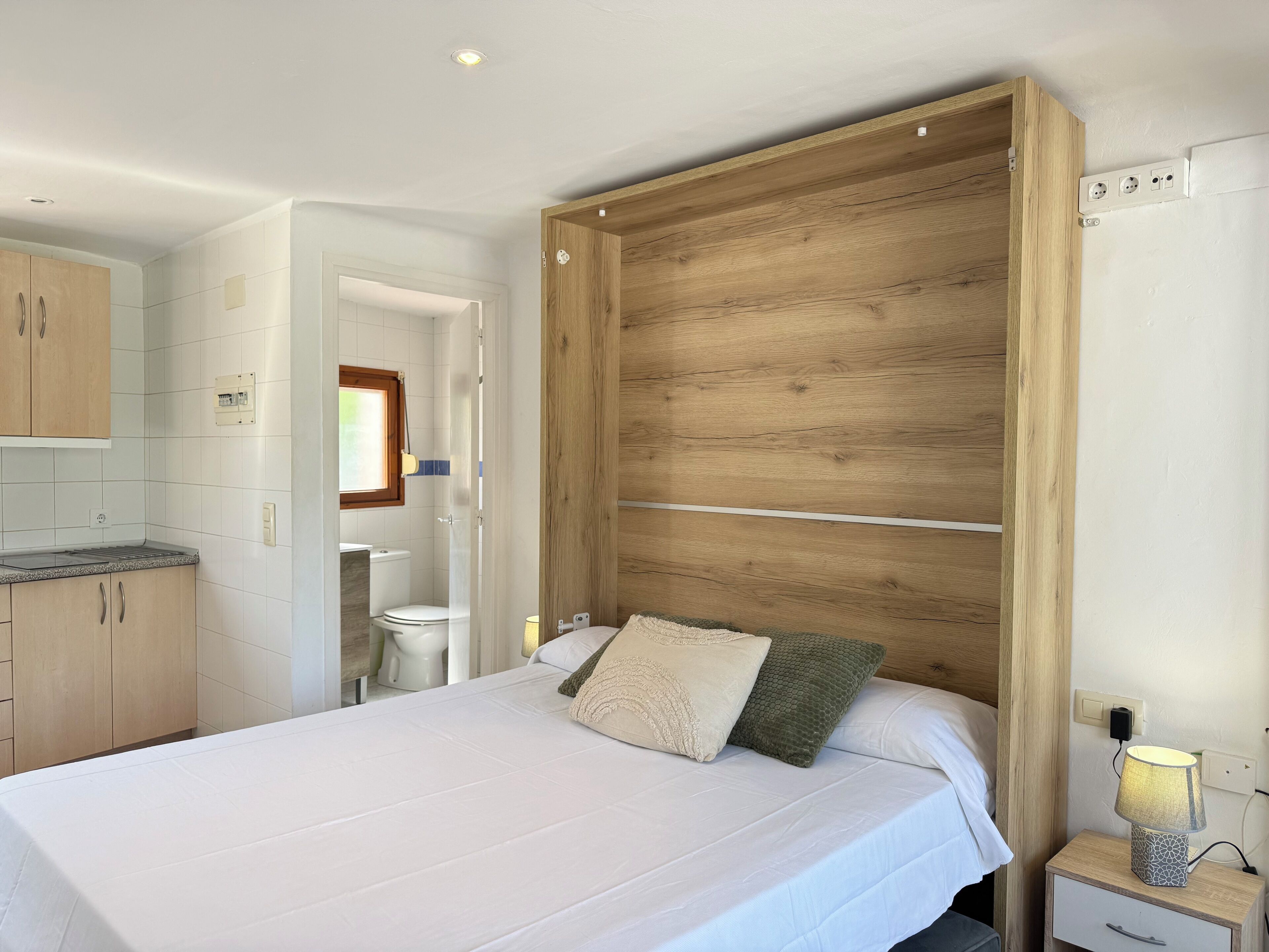 Ein Schlafzimmer mit einem großen Bett, einem Holzkopfteil, einem Nachttisch mit Lampe und Blick ins Badezimmer.