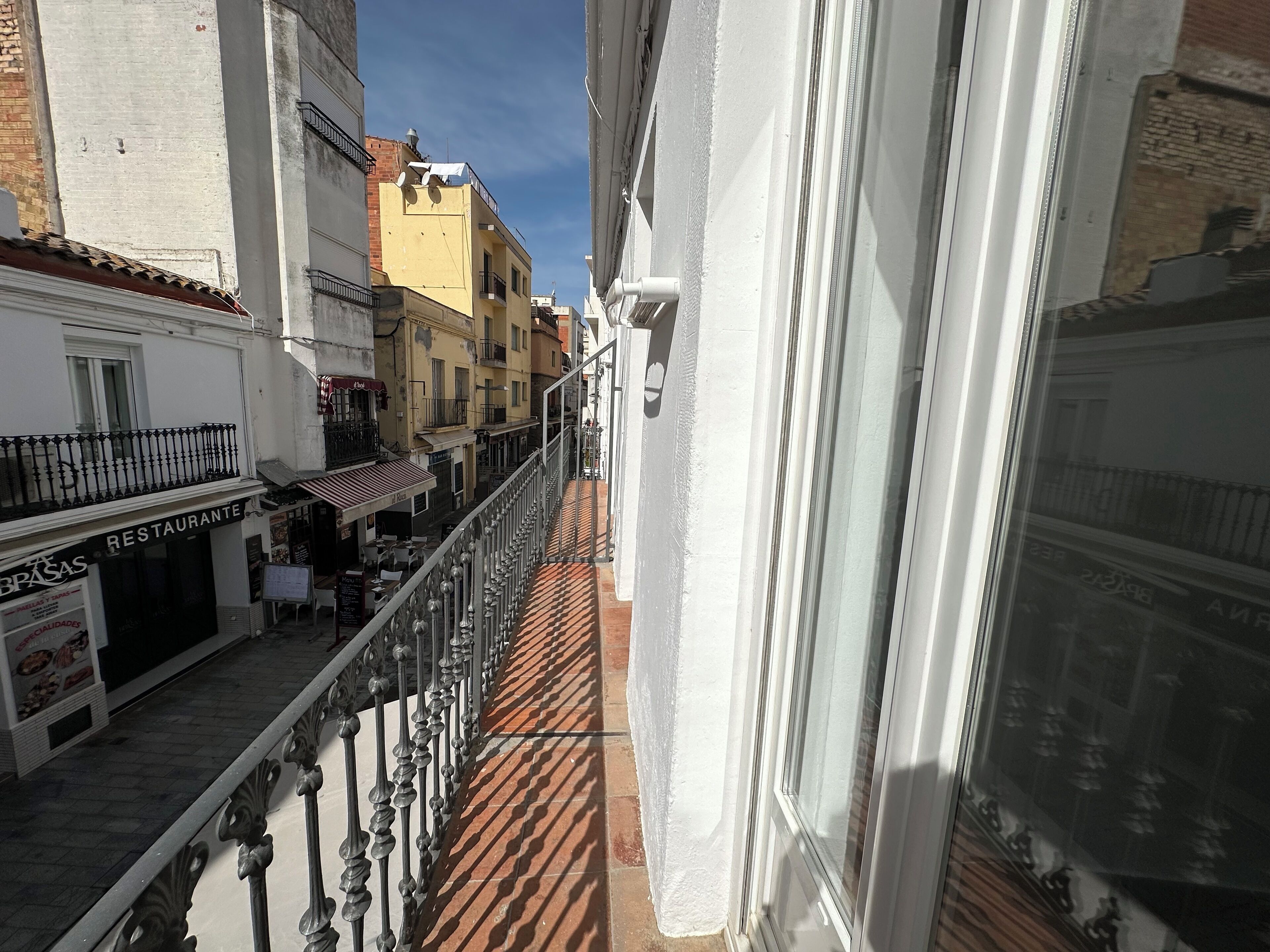 A narrow street with buildings on either side, a balcony with a metal railing, and a restaurant sign.