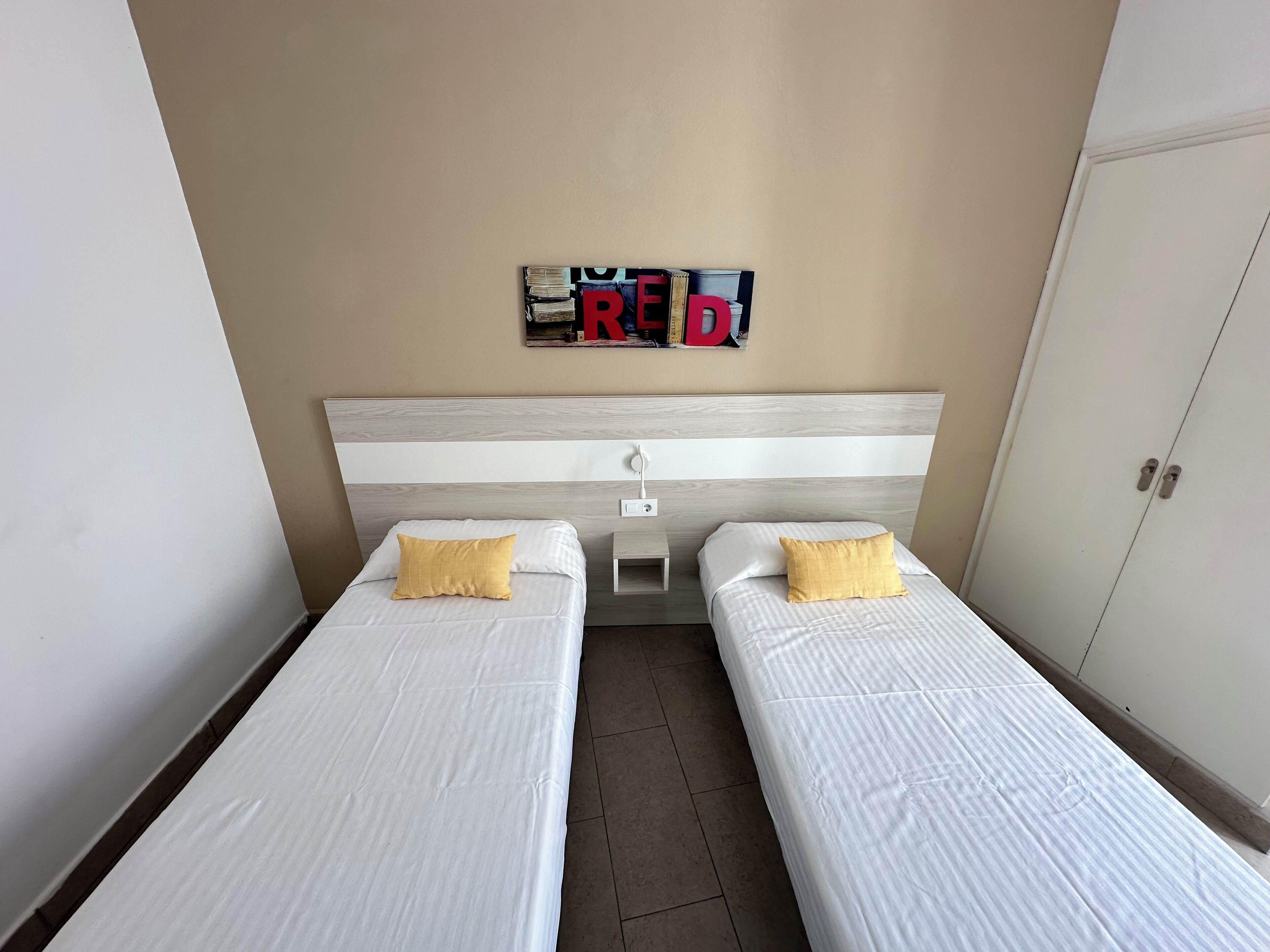 A room with two single beds, a headboard with a light wood finish, and a wall-mounted shelf with books and a red letter 'R'.