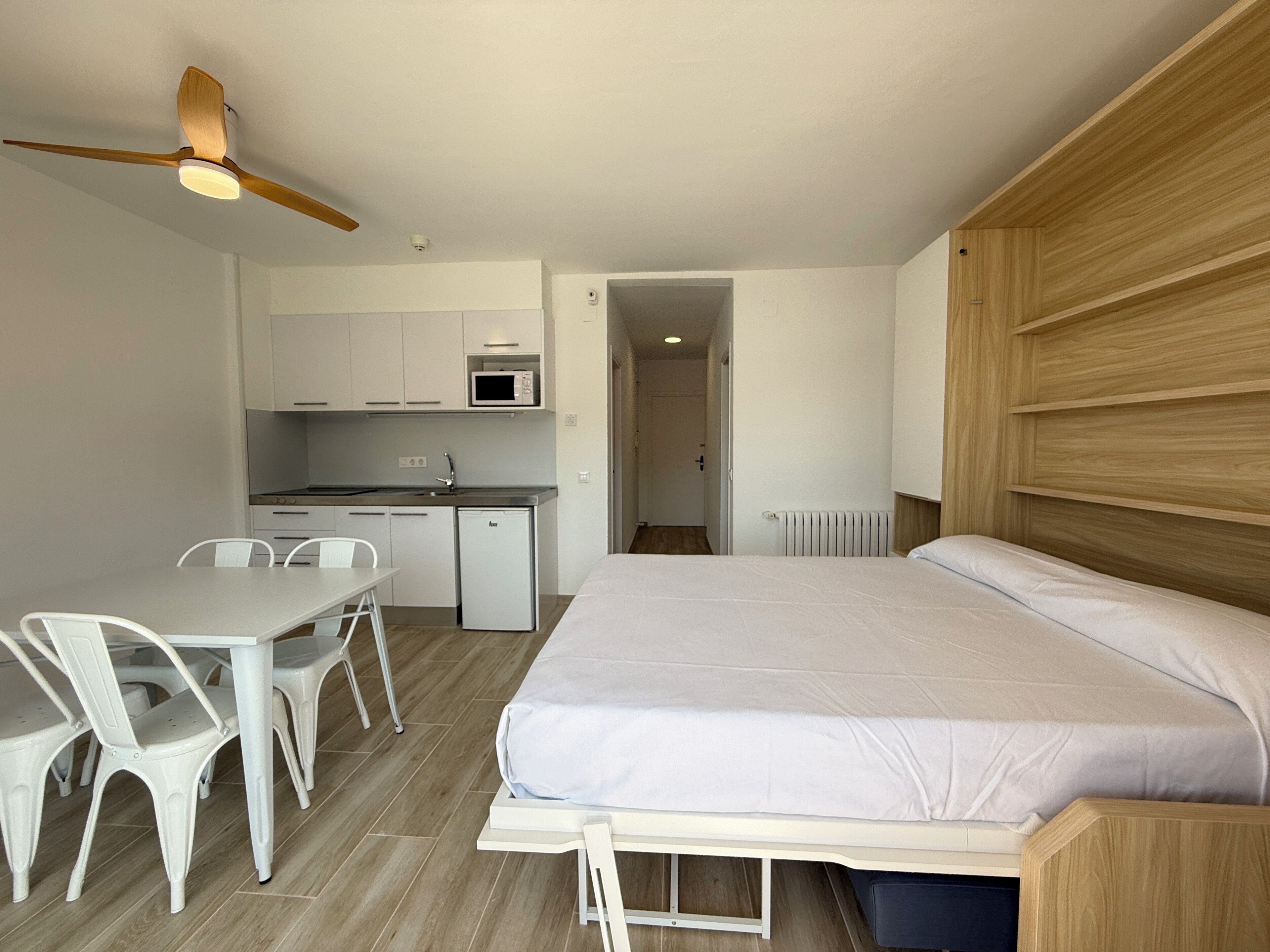 A compact room with a bed, a dining table with chairs, a kitchenette, and a ceiling fan.
