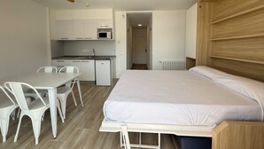 Een compacte kamer met een bed, een eettafel met stoelen, een kitchenette en een plafondventilator.