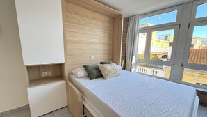 Une chambre à coucher comprenant un lit, une armoire intégrée, une petite étagère et une fenêtre donnant sur des immeubles.