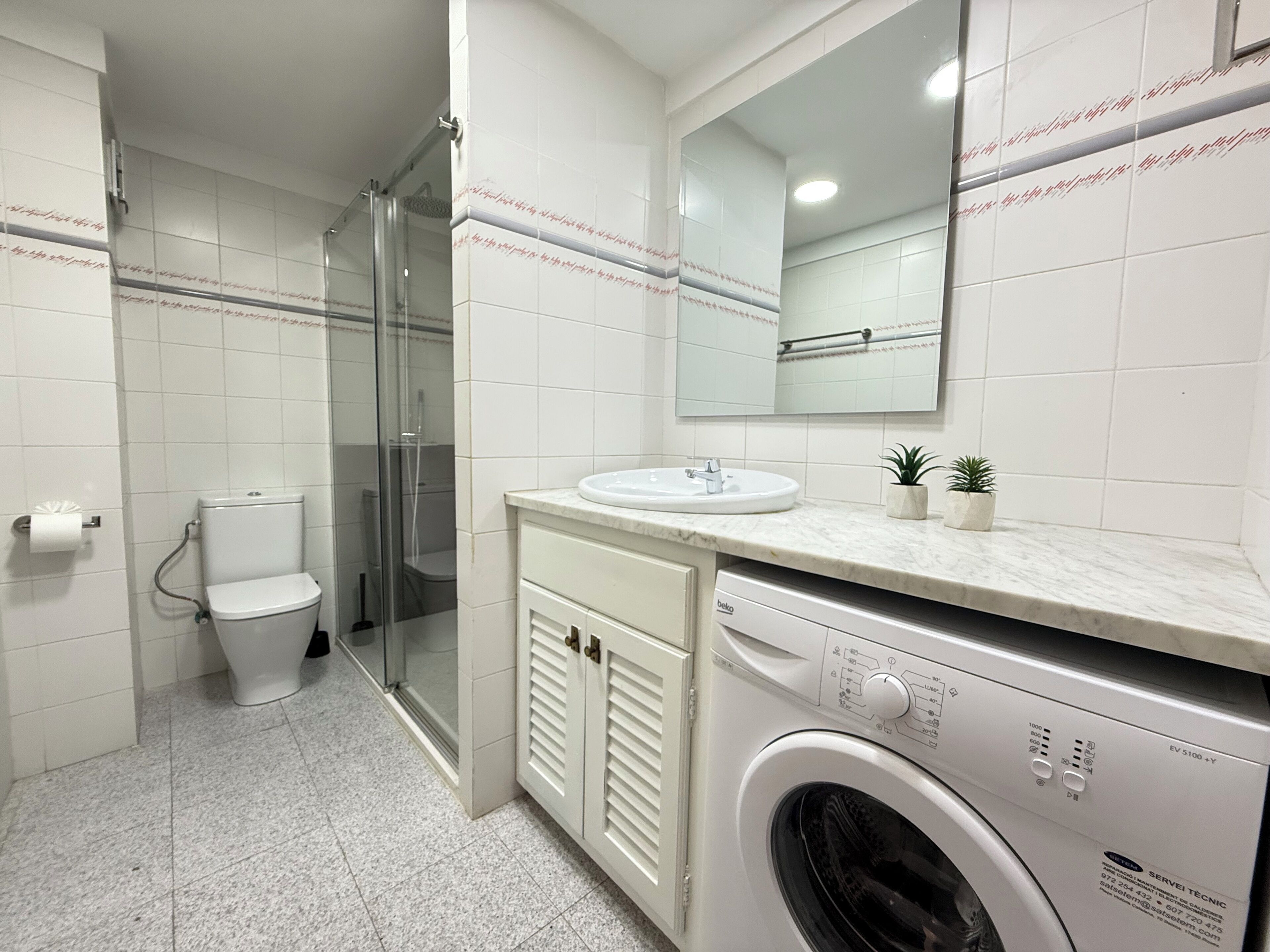 A modern bathroom with a glass-enclosed shower, a toilet, a sink, and a front-loading washing machine.