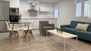 Een moderne keuken met witte kasten, een ingebouwde oven en een roestvrijstalen afzuigkap. Er is een eethoek met een glazen tafel en witte stoelen. In de woonkamer staat een blauwe bank en een klein witte tafeltje.