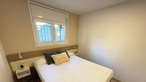 Een slaapkamer met een groot bed, een nachtkastje, een klein tafeltje, een raam met zonwering en een wandlamp.