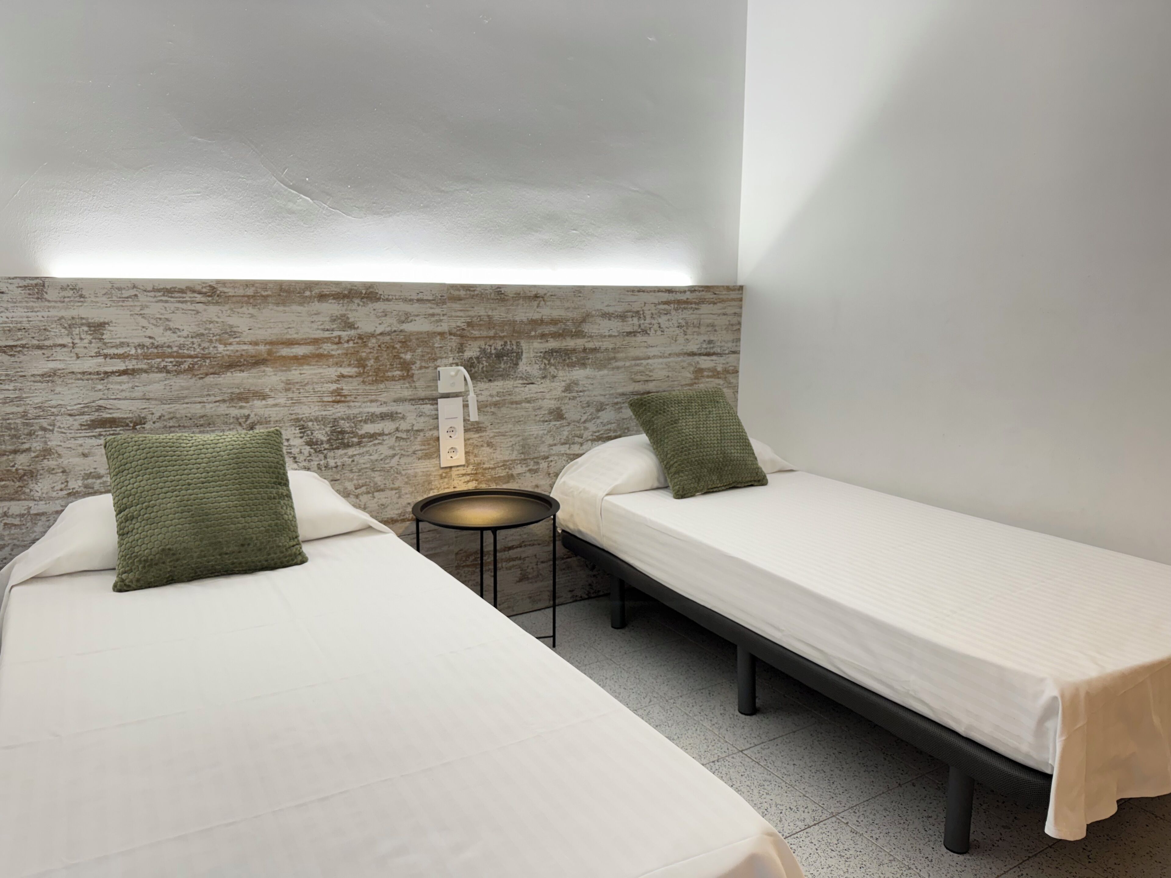 A room with two single beds, each with a green pillow, a small round table, and a wall-mounted light fixture.