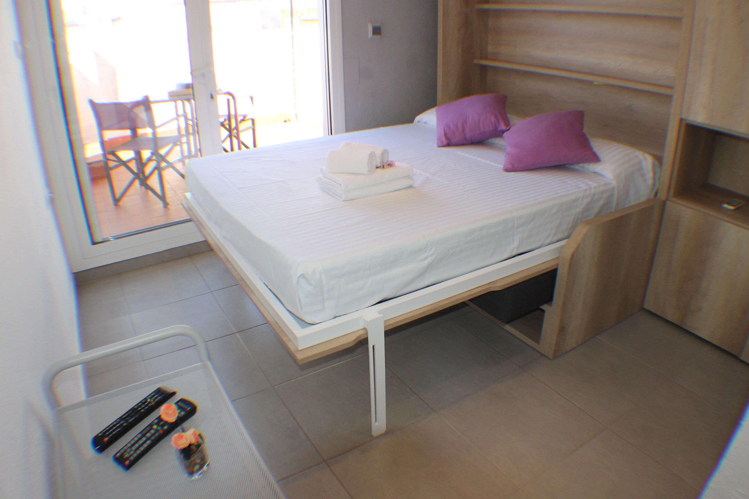 A single bed with a white mattress and two pillows, a small table with a remote control and a glass, and a chair visible through a glass door.