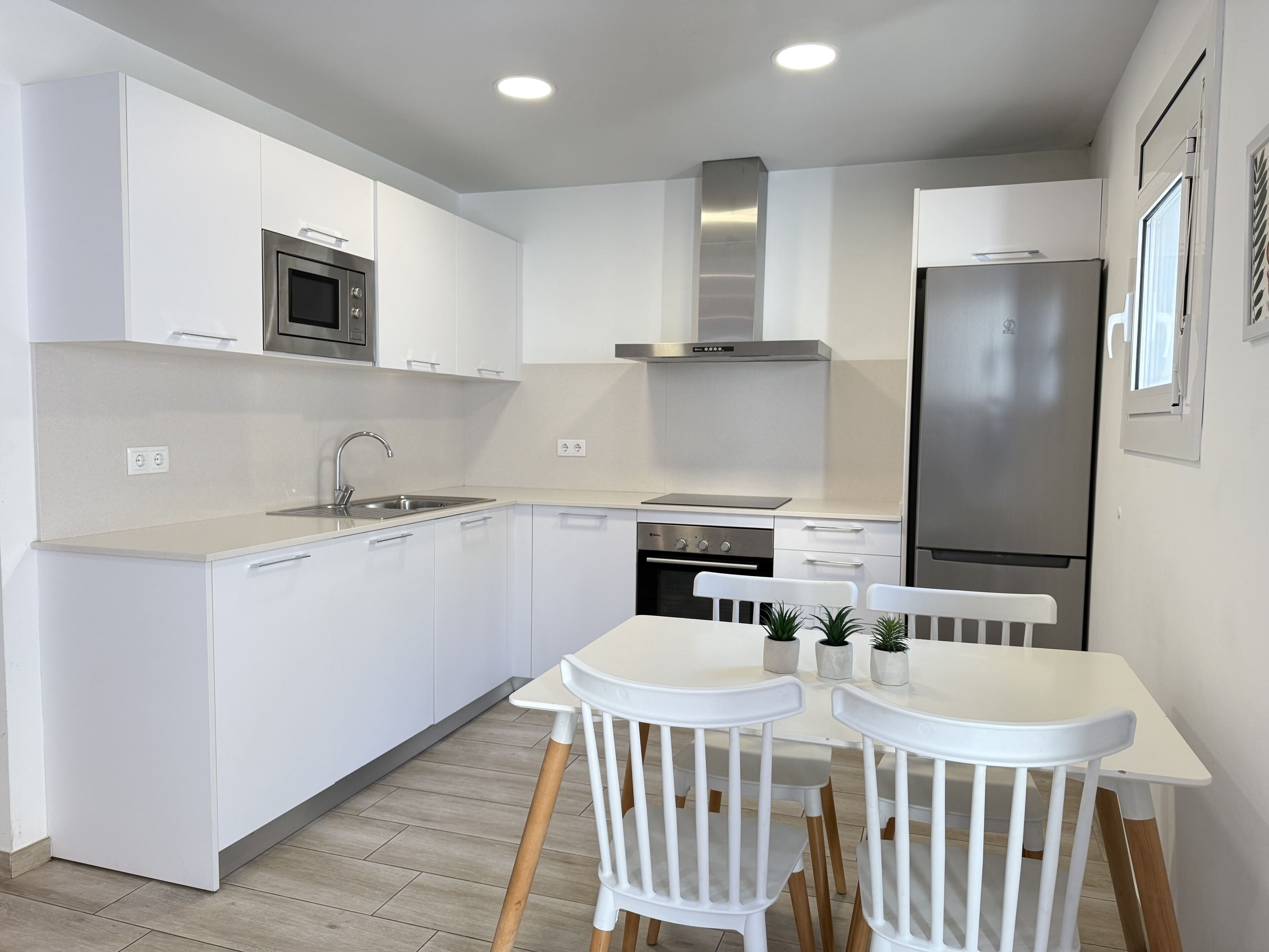 A modern kitchen with white cabinetry, a stainless steel refrigerator, a built-in oven, and a microwave. There is a dining table with two chairs and a small potted plant on it.