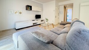 En moderne stue med en grå hjørnesofa, et fladskærms-tv, et hvidt tv-møbel og et lille bord med en plante.