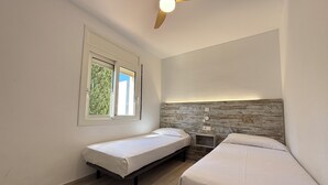 Een kamer met twee eenpersoonsbedden, een plafondventilator, een raam met uitzicht op groen en een nachtkastje dat aan de muur bevestigd is.