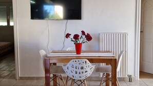 Ruokailutilassa on puinen pöytä, valkoiset tuolit ja punainen maljakko, jossa on kukkia. Seinälle, pöydän yläpuolelle, on asennettu litteänäyttöinen televisio.