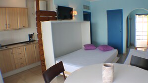 Una habitación compacta con cama, una mesita, una silla, televisor y kitchenette.