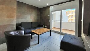 Un salon moderne avec un canapé, une table basse et un petit pouf. Une porte-fenêtre coulissante donne accès à un balcon d’où l’on peut admirer la vue sur les immeubles.