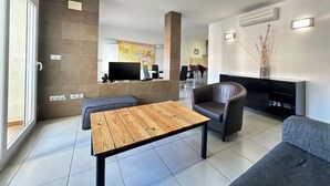 Un salon moderne avec une table basse en bois, un fauteuil en cuir et un canapé gris. Un téléviseur à écran plat est fixé au mur, et un tableau est visible au fond.