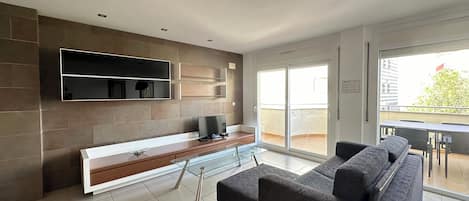 Uma sala de estar moderna com um amplo sofá modular, uma televisão de tela plana fixada na parede, uma mesa de centro de vidro e uma porta de correr de vidro que dá para uma varanda externa.