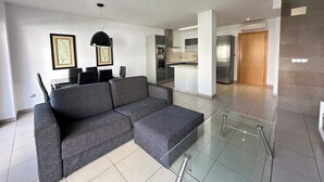 A modern living room with a grey sectional sofa, a dining table with chairs, and a kitchen area with a refrigerator and stove.