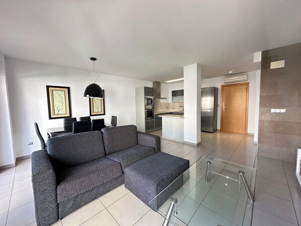 A modern living room with a grey sectional sofa, a dining table with chairs, and a kitchen area with a refrigerator and stove. - Agi Drugstore AE Apartamento for rent in Roses Girona. (Roses)