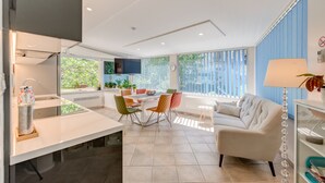 A modern kitchen with a central island, a dining area with colorful chairs, a sofa, and a large window with a view of greenery. - Luxury apartment positioned in a green oasis in the middle of the city (Split)