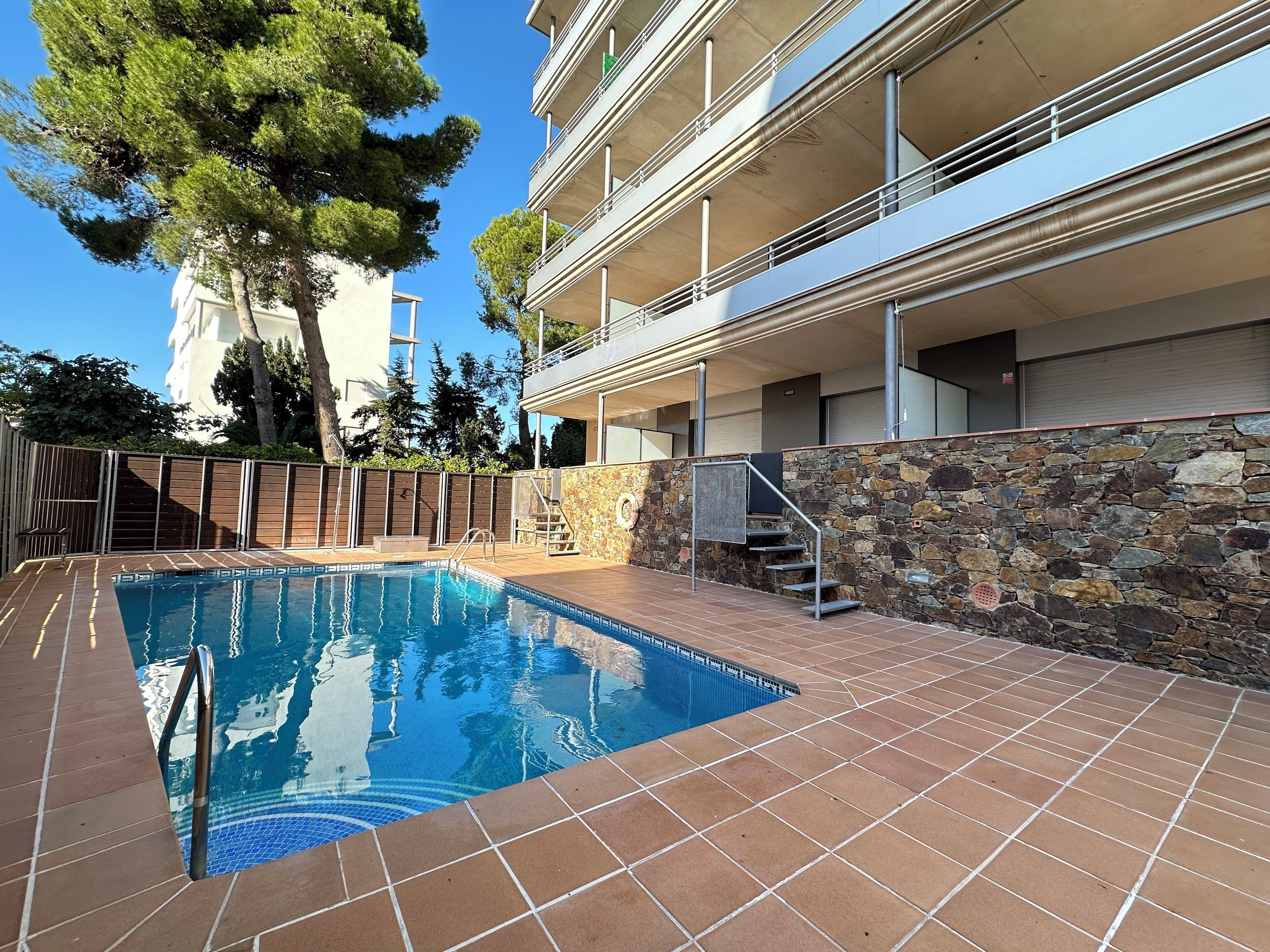 A swimming pool with a stone wall and metal railing, surrounded by a tiled deck and a stone wall.