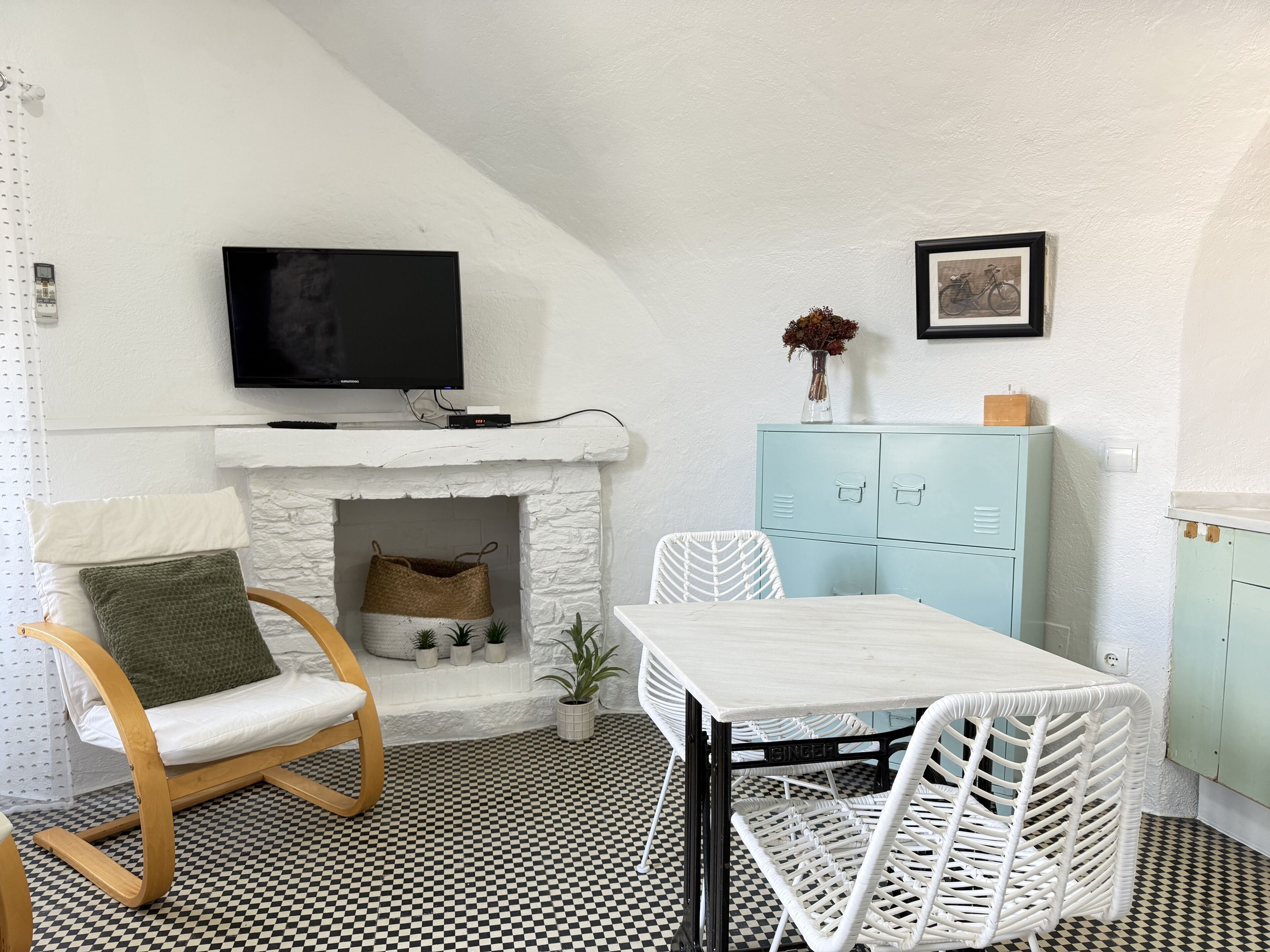 A room with a checkered floor, a white table with chairs, a wooden chair with a cushion, a white fireplace, a television on the wall, and a light blue cabinet.