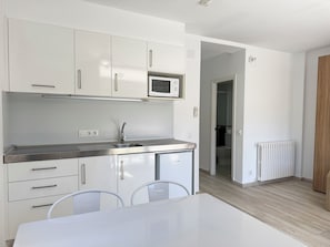 Uma cozinha moderna com armários brancos, uma pia de aço inoxidável e um micro-ondas. Há uma mesa de jantar com cadeiras.
