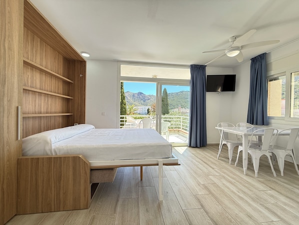 Quarto com cama, guarda-roupa de madeira, varanda com cadeiras e televisão.