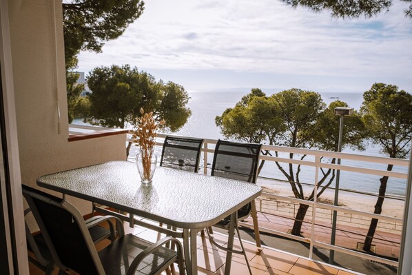 A balcony with a table and chairs, overlooking a beach with trees and a clear sky.