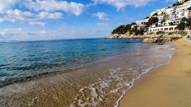 Ein Strand mit klarem Wasser, sandigem Ufer und Gebäuden an einem Hügelhang.