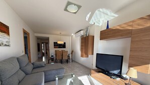 A modern living room with a grey sectional sofa, a glass coffee table, a wooden TV stand, and a dining area in the background. - Agi Riera Ginjolers 3B Apartamento for rent in Roses Girona. (Roses)