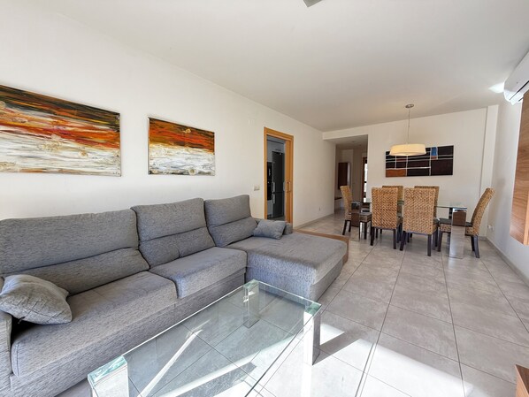 A modern living room with a grey sectional sofa, a glass coffee table, and a dining area with wicker chairs and a glass table. - Agi Riera Ginjolers 3B Apartamento for rent in Roses Girona. (Roses)