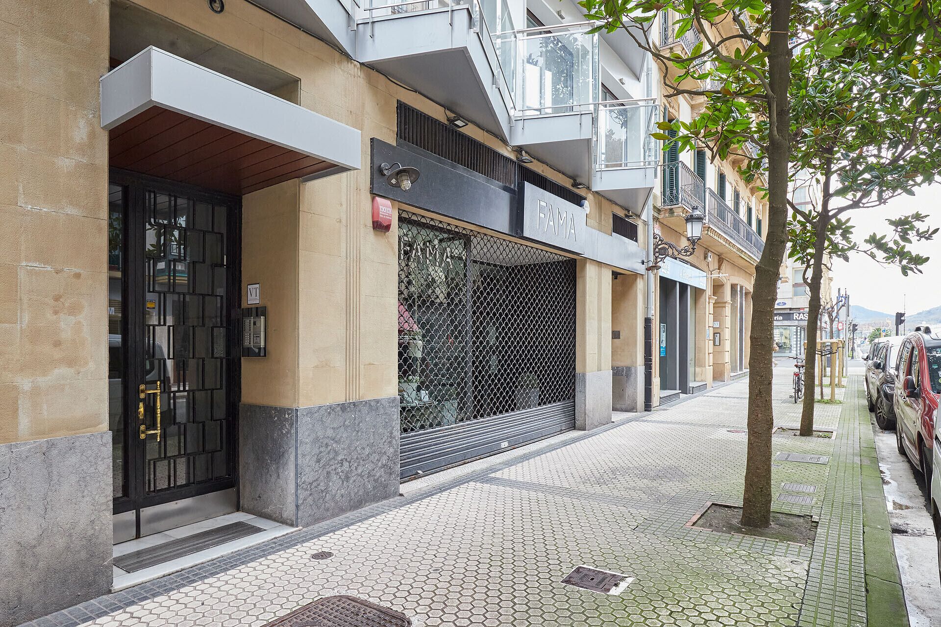 A building with a black metal gate, a visible 'FAMA' sign, and a tiled sidewalk.