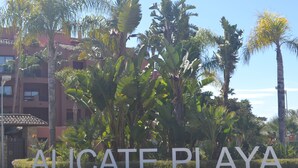 A sign with the text "ALICATE PLAYA" surrounded by lush greenery and palm trees. - URBANIZATION ALICATE PLAYA. (Marbella)